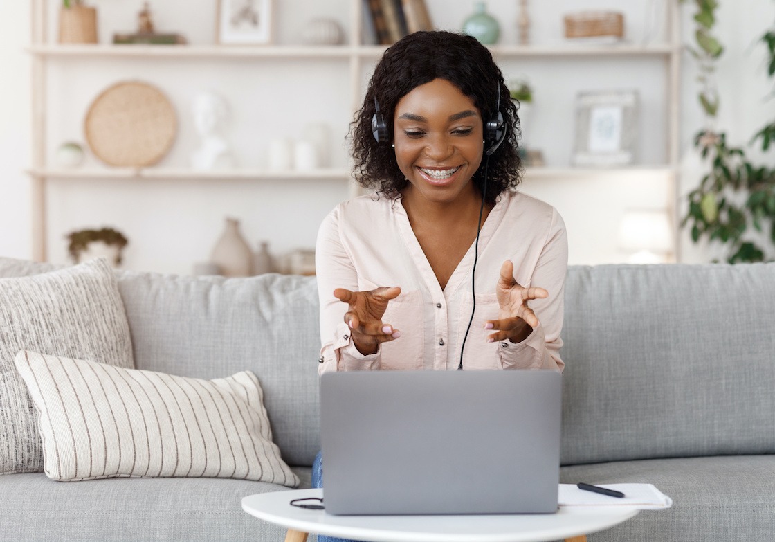 Online Tutoring Concept. Smiling black female tutor having video call with student, giving language class by webcam, sitting on couch at home