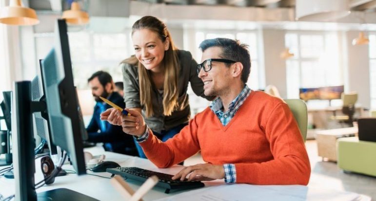 Two coworkers collaborating at a computer during a meeting, reviewing work and smiling in a modern office